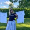 Two white T-shirts are hanging and woman shows the message of the black T-shirt that she is wearing.