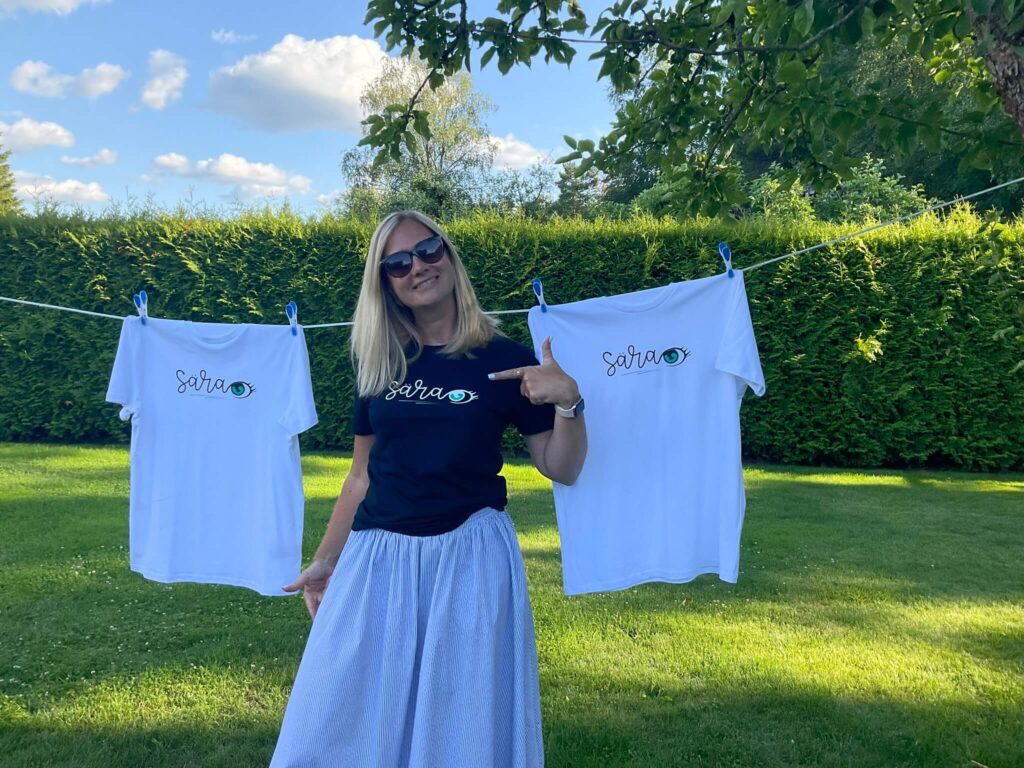 Two white T-shirts are hanging and woman shows the message of the black T-shirt that she is wearing.